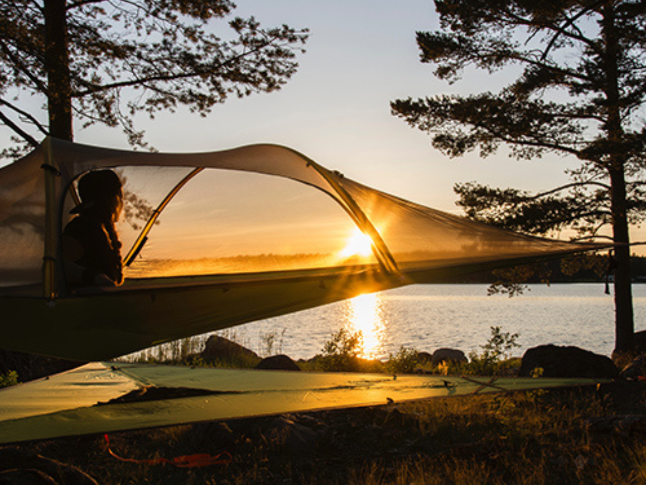 Bild på en person sittandes i ett trädtält i en solnedgång intill en sjö. 