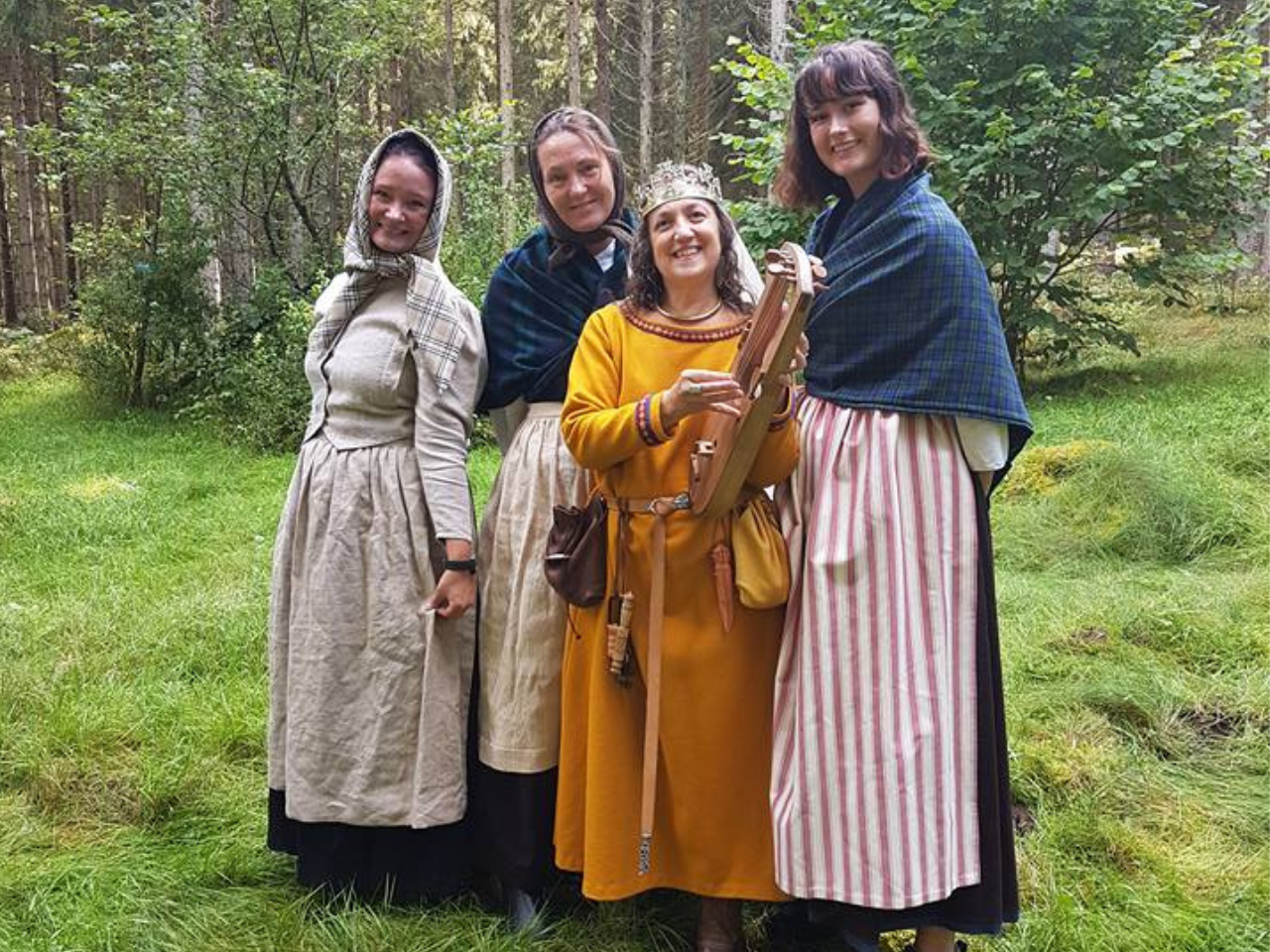 Fyra kvinnor i medeltidskläder står i en skogsglänta. En av kvinnorna håller i ett stränginstrument.