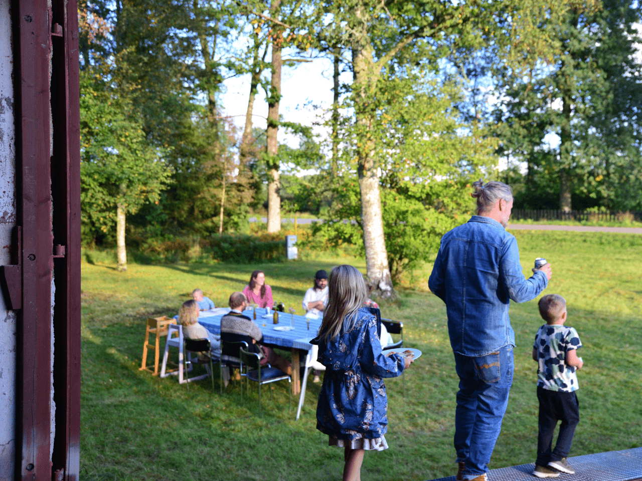 Man och två barn på väg ut i en trädgård bärandes på fika. Picnicbord i bakgrunden.