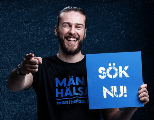 Bild på en man som håller upp en skylt med texten "Sök nu!". På mannens tröja står det "Män i hälsa".